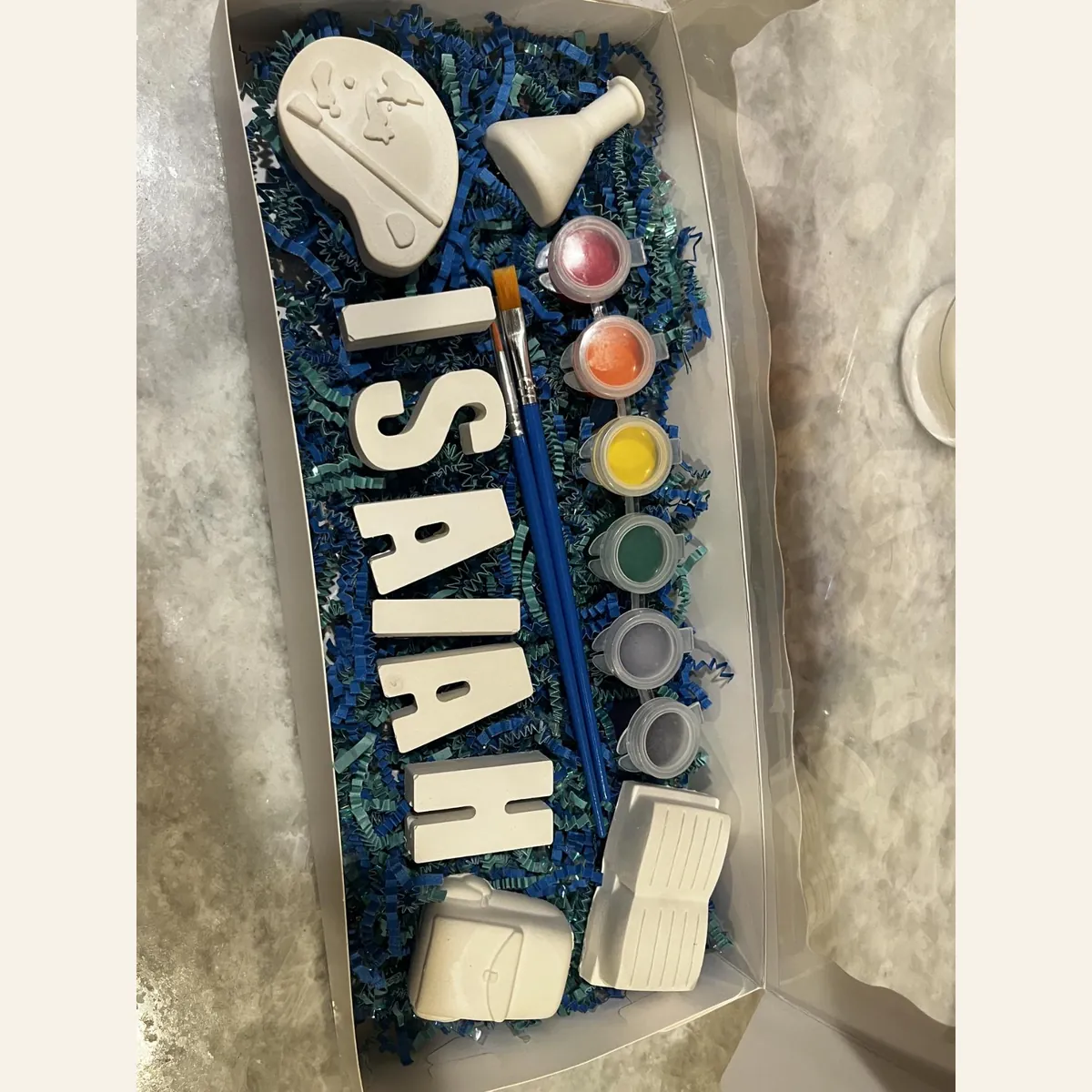 Finished name kit in a box, with plaster letters spelling Isaiah, a paint palette, a bottle, two brushes, six paint pots, and two small plaster pieces